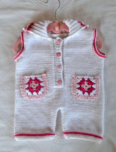 Weißer gestrickter Babystrampler mit rosa Granny‑Square‑Taschen und rosa Knöpfen an einem rosafarbenen Kleiderbügel