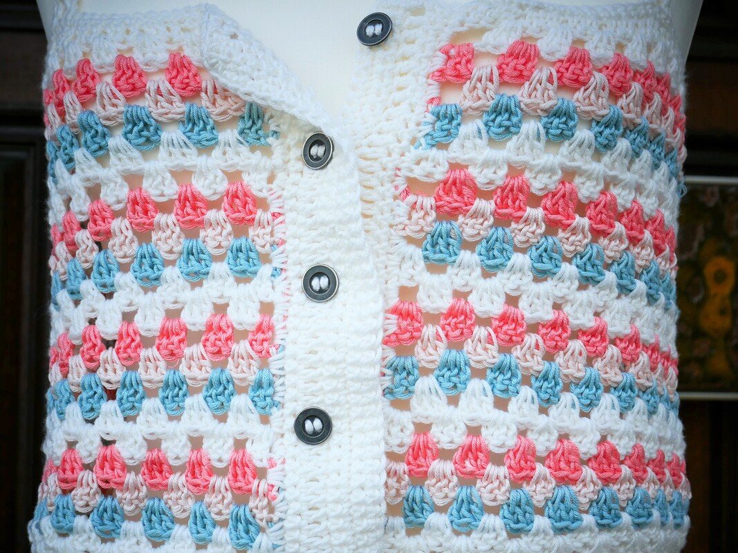 Nahaufnahme einer handgehäkelten Damenweste im Granny-Muster in Weiß, Rosa und Hellblau mit sichtbarer Knopfleiste