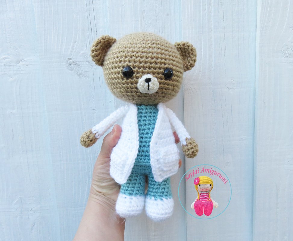 Brown crocheted amigurumi teddy bear wearing a white coat and teal scrubs, held by a hand.
