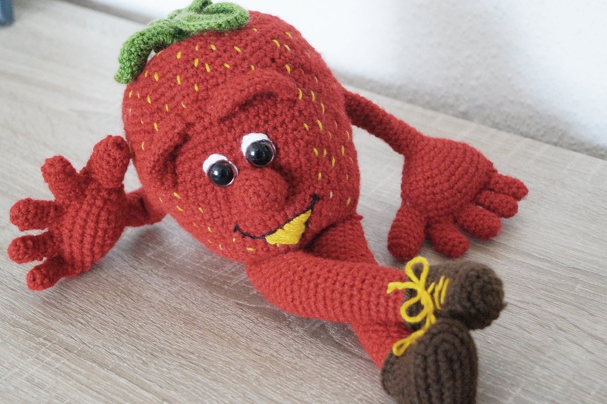 Handgehäkelte rote Erdbeerfigur (Amigurumi) mit Gesicht, Armen und braunen Schuhen auf hellem Holz