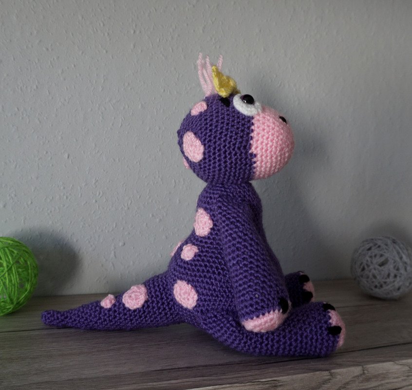 Gehäkelter lila Amigurumi-Drache mit rosa Punkten und gelbem Horn auf Holzoberfläche