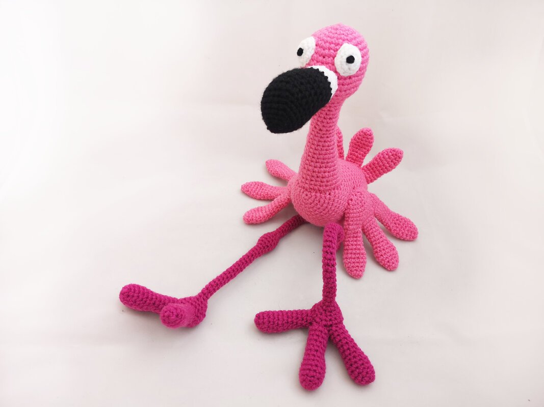 Pink crocheted flamingo amigurumi toy with a black beak and white eye details on a white background.