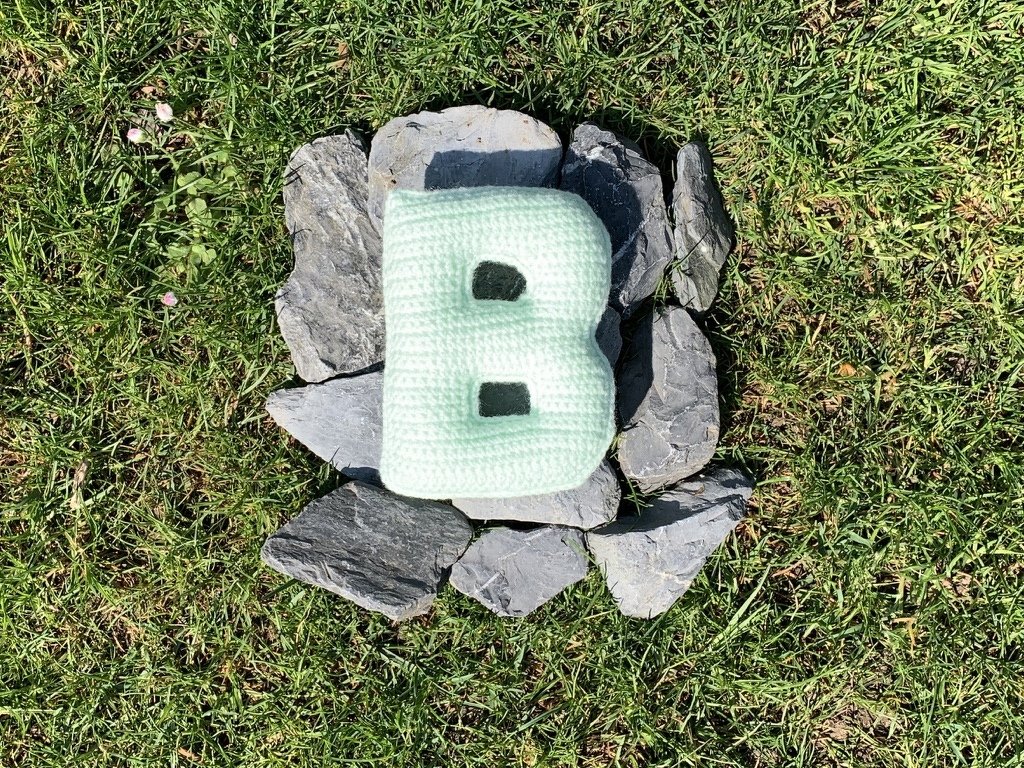 Gehäkeltes hellmintfarbenes Kissen in Form des Buchstabens B auf Schiefersteinen im Rasen
