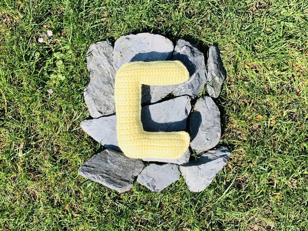 Gehäkelter gelber Buchstabe C auf grauen Steinen im Gras