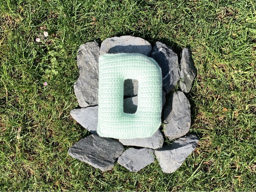 Nahaufnahme eines gefüllten, gehäkelten Kissens in Form des Buchstabens D auf Schiefersteinen