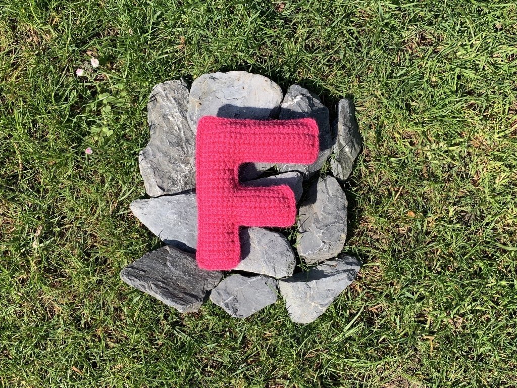 Gehäkelter Buchstabe F in Pink auf Grausteinen im grünen Rasen