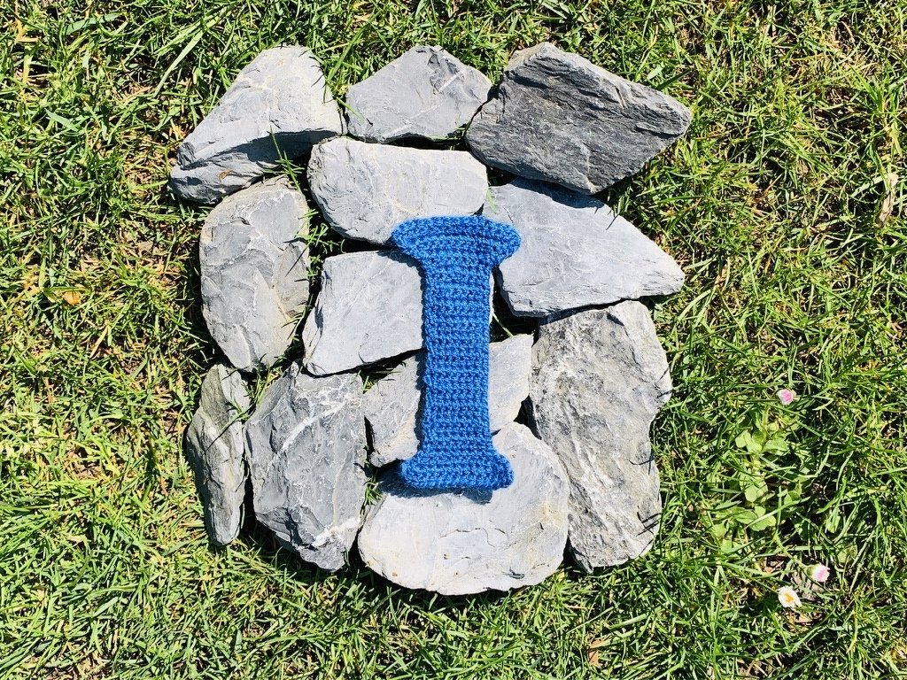 Gehäkelter blauer Buchstabe I auf grauen Steinen im Gras