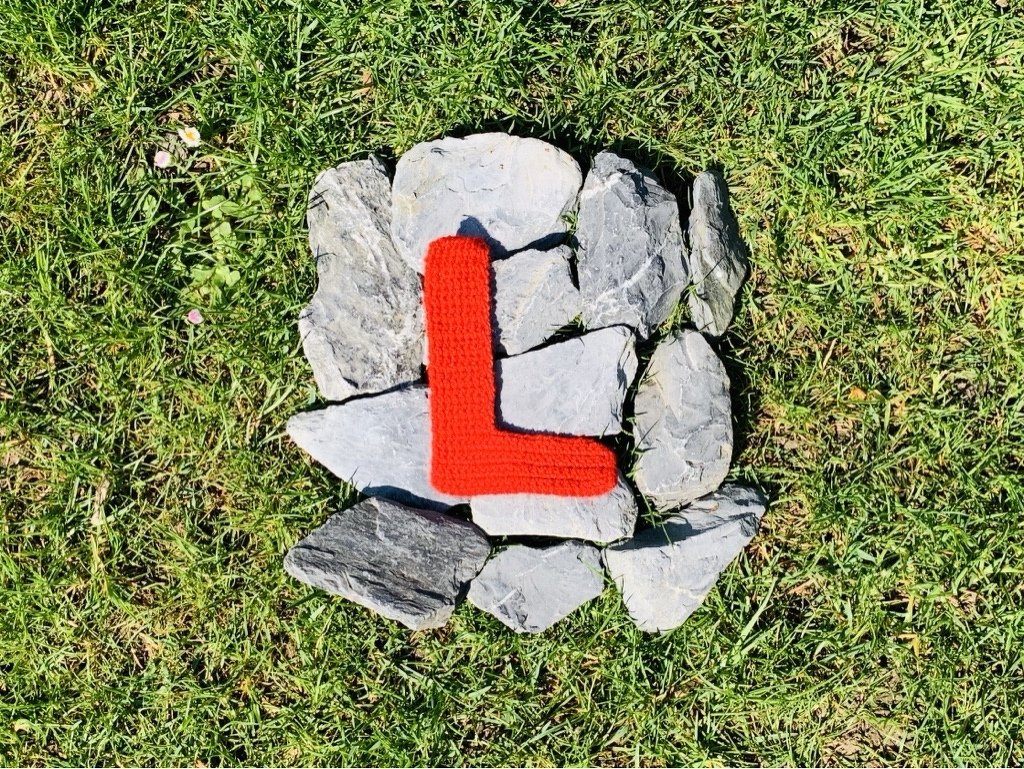 Roter gehäkelter Buchstabe L auf grauen Steinen im Gras