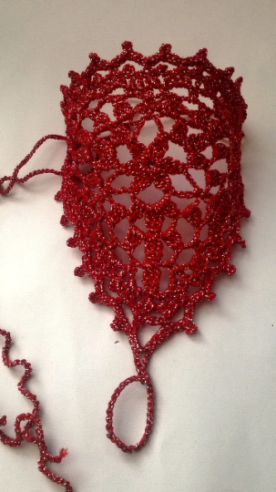 Red crocheted barefoot sandal with glittering thread, openwork lace pattern and a toe loop on a plain light background.