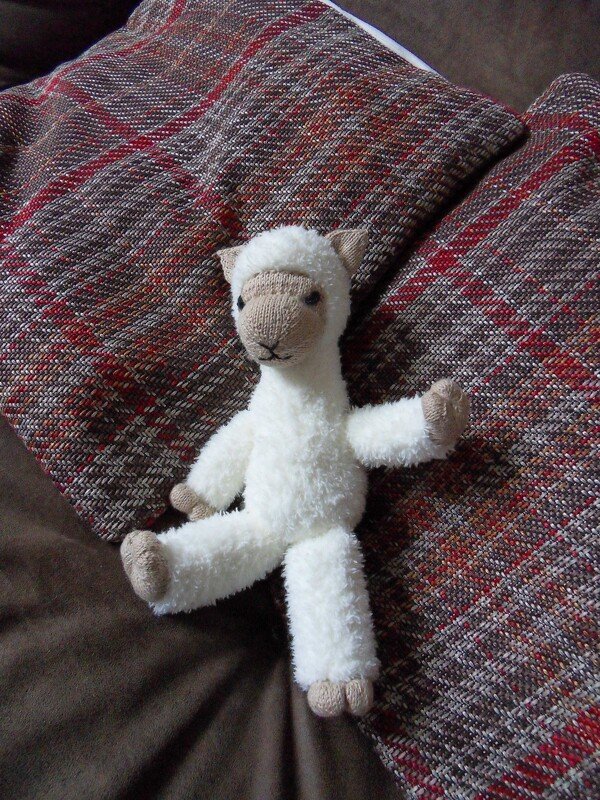 Gestricktes weißes Lamm-Amigurumi mit beigem Gesicht und Pfoten auf rot-brauner Decke