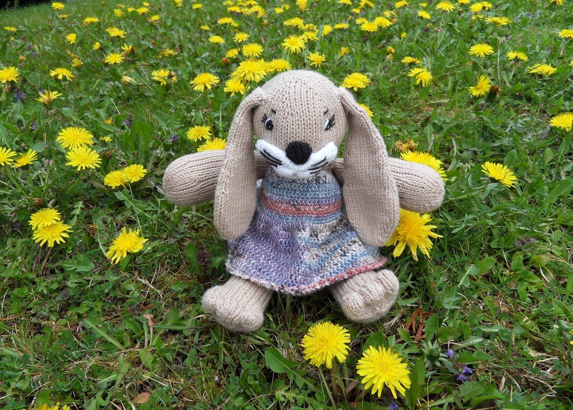 Gestrickter Amigurumi-Hase in Hellbraun mit Schlappohren und meliertem Strickkleid im Gras.
