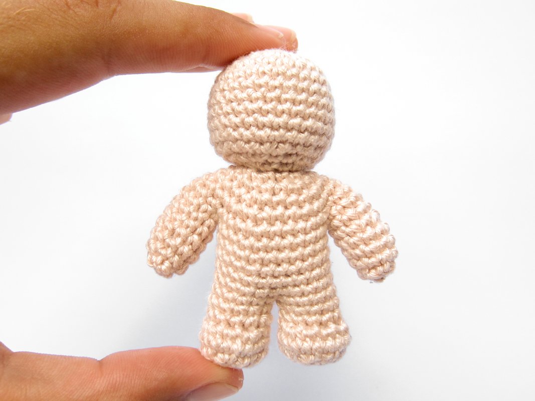 Close-up of a small, light beige crocheted amigurumi doll body held between fingers against a white background.