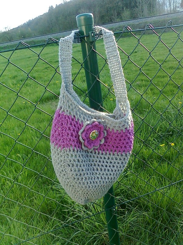 Gehäkelte graue Tasche mit pinkem Streifen und gehäkelter Blüte hängt an einem Zaun