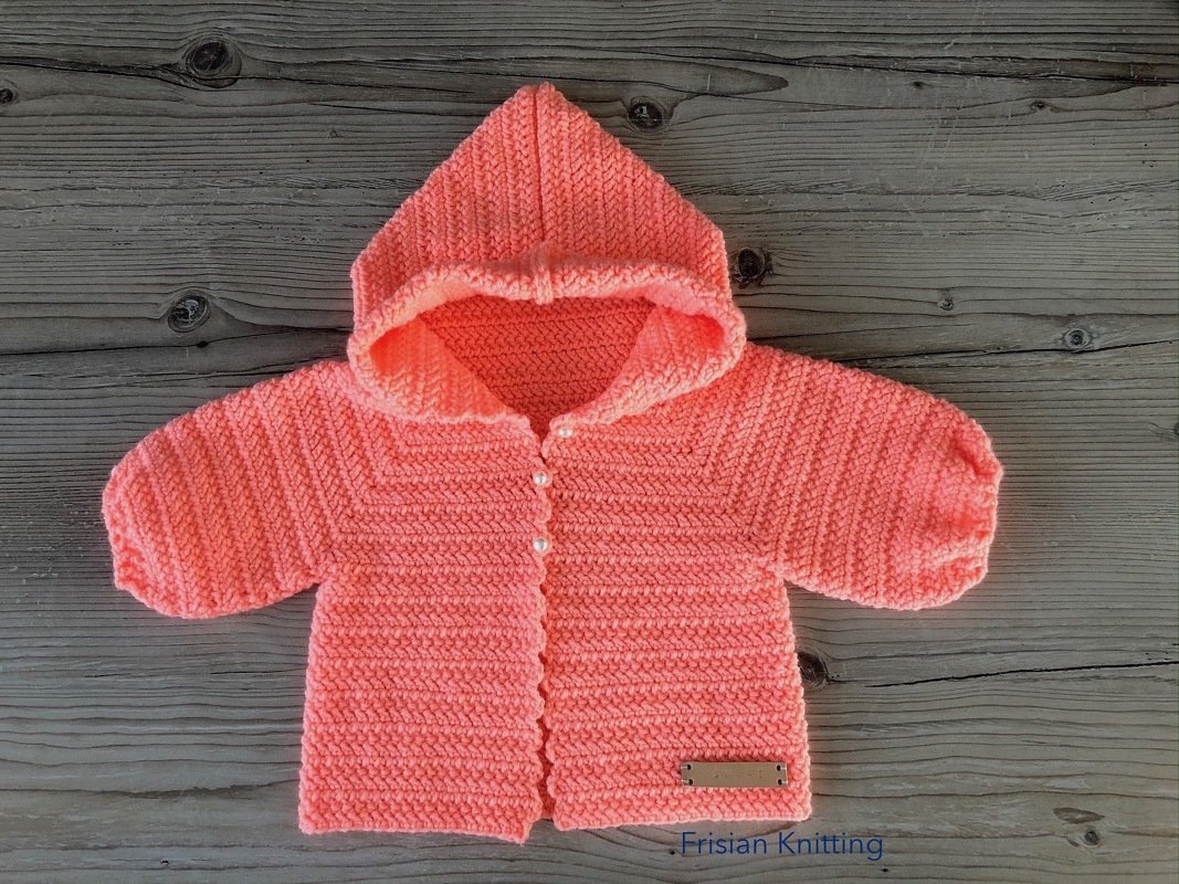 Gehäkelte Kapuzenjacke für Babys in Koralle mit Perlmuttknöpfen auf Holzhintergrund.