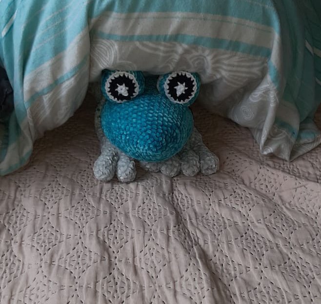 Verspieltes gehäkeltes Froschkissen in Blau und Türkis mit auffälligen Augen, auf einem Bett platziert