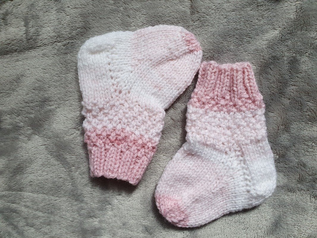 Zwei gestrickte Babysöckchen in Rosa und Weiß mit Rippbündchen auf grauem Stoff