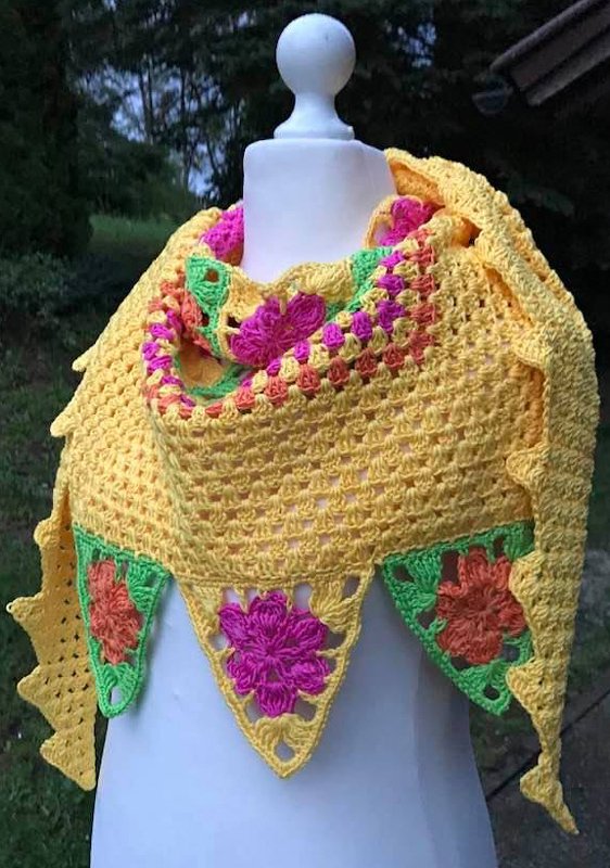Gehäkeltes gelbes Dreieckstuch mit pinken, orangen und grünen Blumenmotiven auf einer Schneiderbüste