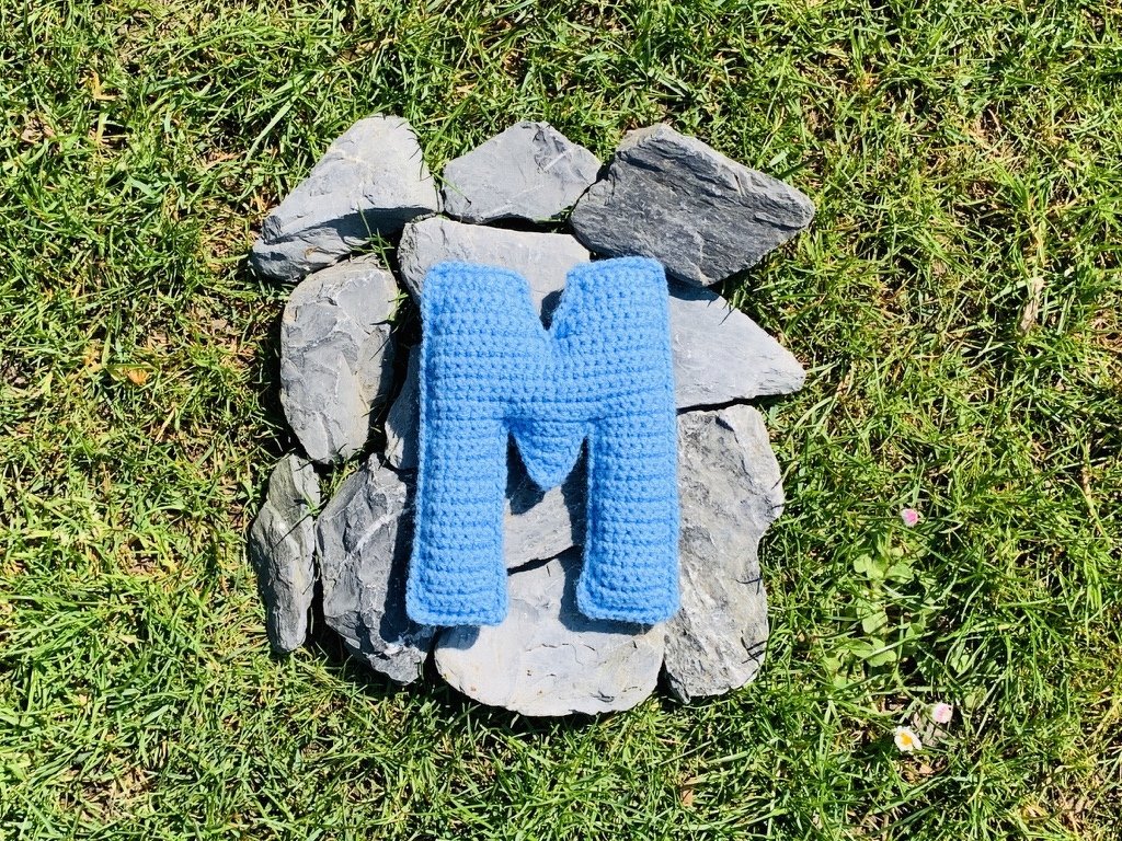 Gehäkelter blauer Buchstabe M auf grauen Steinen in grünem Gras