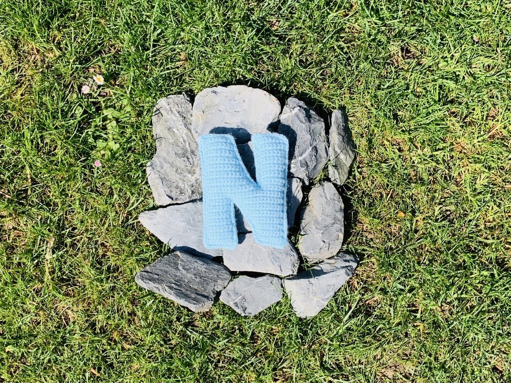 Hellblauer gehäkelter Buchstabe N auf grauen Schieferplatten im Gras