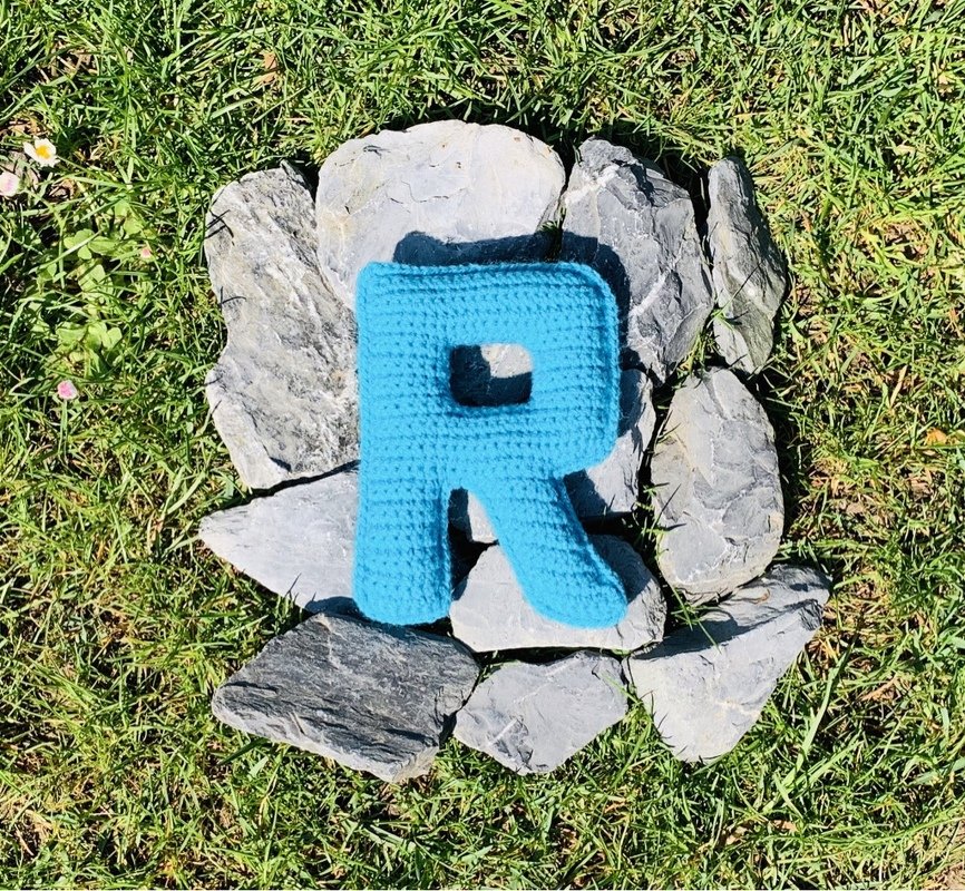 Gehäkelter Buchstabe R (türkis) auf Gras, umgeben von Steinen