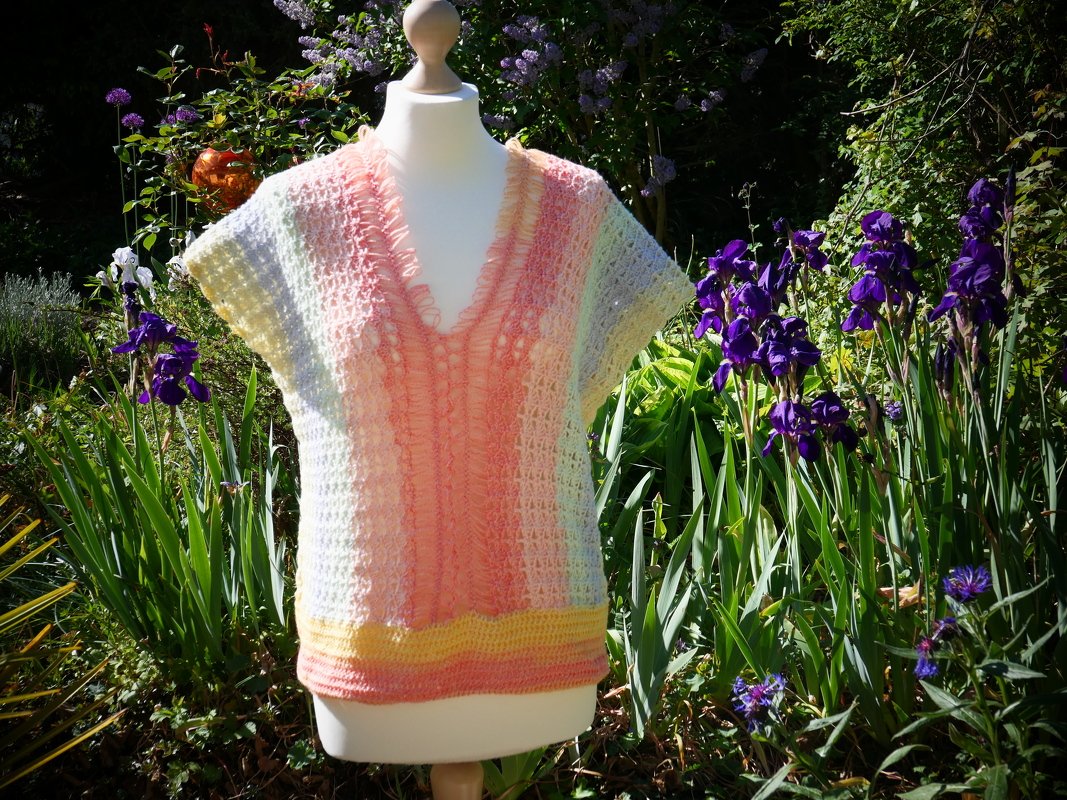 Gehäkeltes Pastell-Damen-Shirt mit V-Ausschnitt auf Schneiderpuppe im Garten bei lilanen Irisblüten.