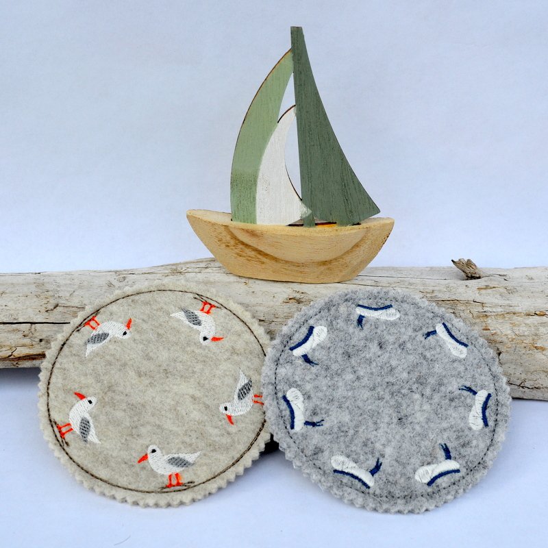 Zwei runde Filz‑Tassenteppiche, einer hellgrau mit Möwen, einer grau mit Wellen, auf Treibholz neben einem Holzsegelboot.