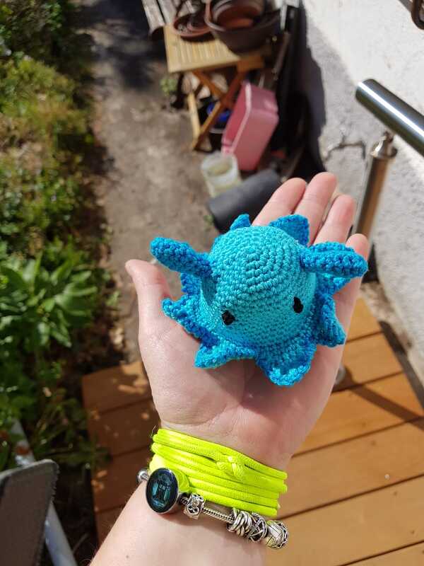 Hand mit neon-gelbem Armband hält einen kleinen türkisfarbenen gehäkelten Axolotl mit schwarzen Augen im Freien