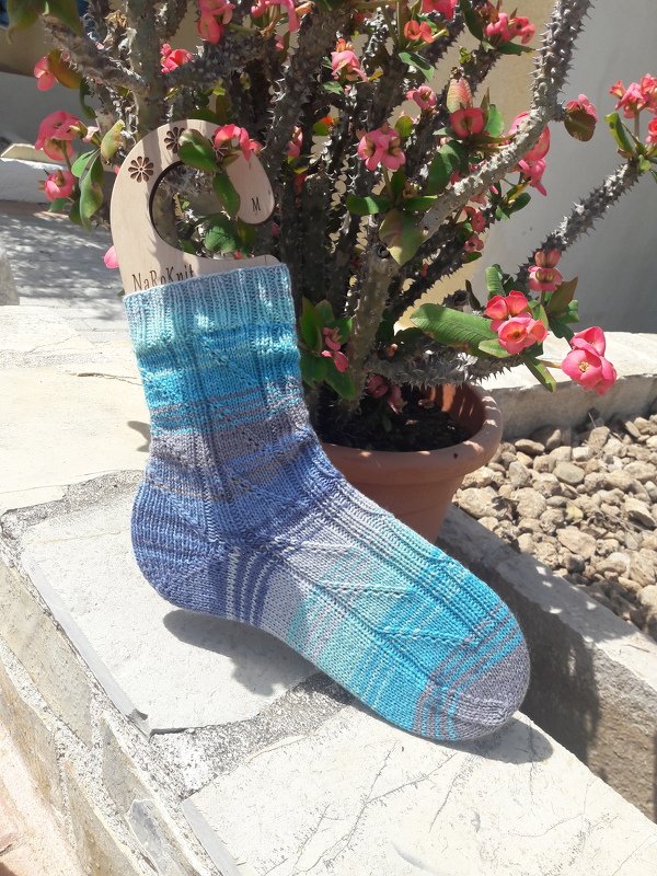 Gestrickte Socke in Blau‑Türkis‑Grau auf einem Sockenbrett vor einer blühenden Topfpflanze.