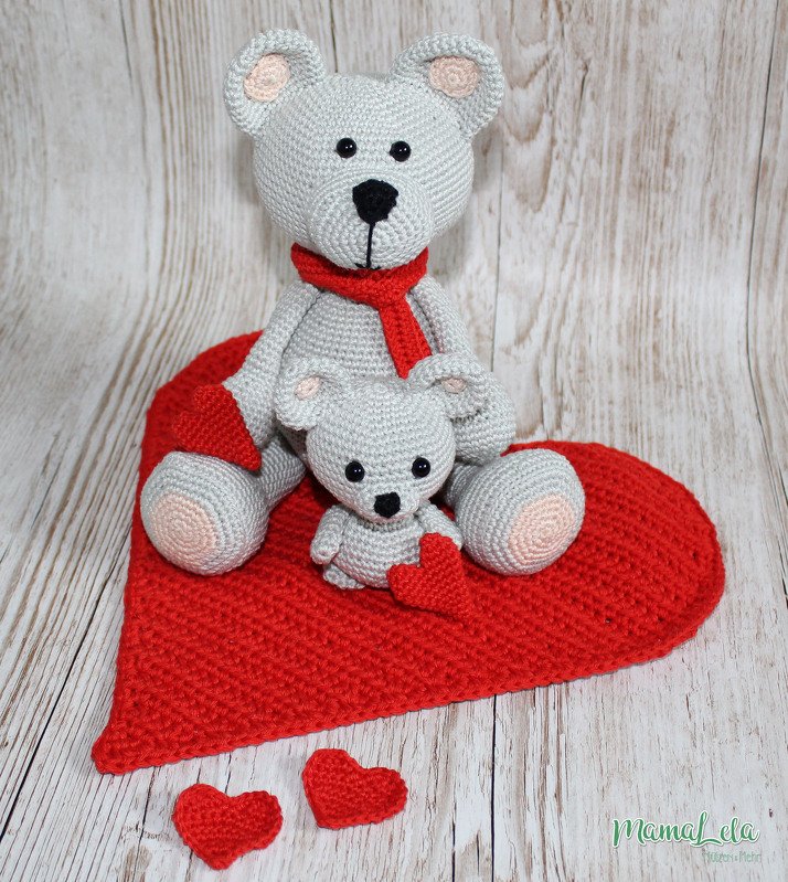 Zwei gehäkelte graue Amigurumi-Teddys auf einem großen roten Häkelherz, mit rotem Schal und kleinen Häkelherzen auf hellem Holzhintergrund.