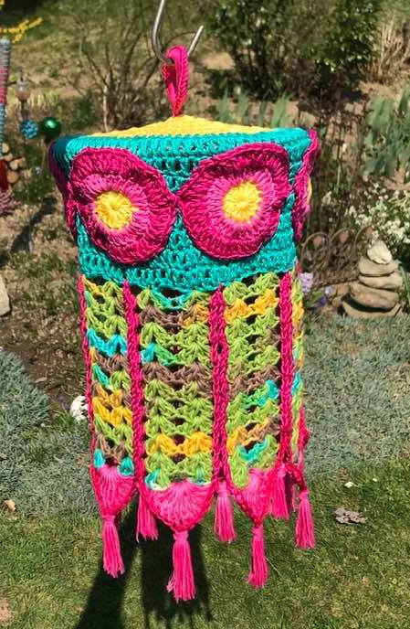 Gehäkelter Lampion in Türkis, Pink und Gelb mit großen Blumen und langen Fransen, hängend im Freien vor grünem Hintergrund.