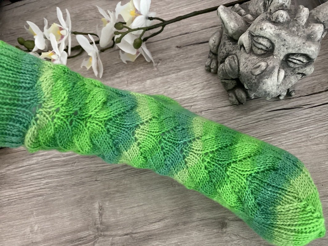 Grüne gestrickte Socke mit Zopfmuster auf hellem Holzhintergrund neben weißen Orchideenblüten und einer steinernen Figur