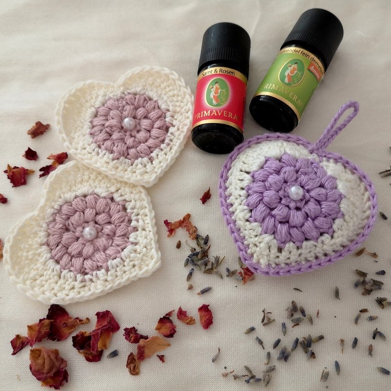 Drei gehäkelte Herzen in Cremeweiß, Rosa und Flieder mit zentralen Perlen auf hellem Stoff, umgeben von getrockneten Rosenblättern, Lavendel und zwei Duftöl‑Fläschchen.