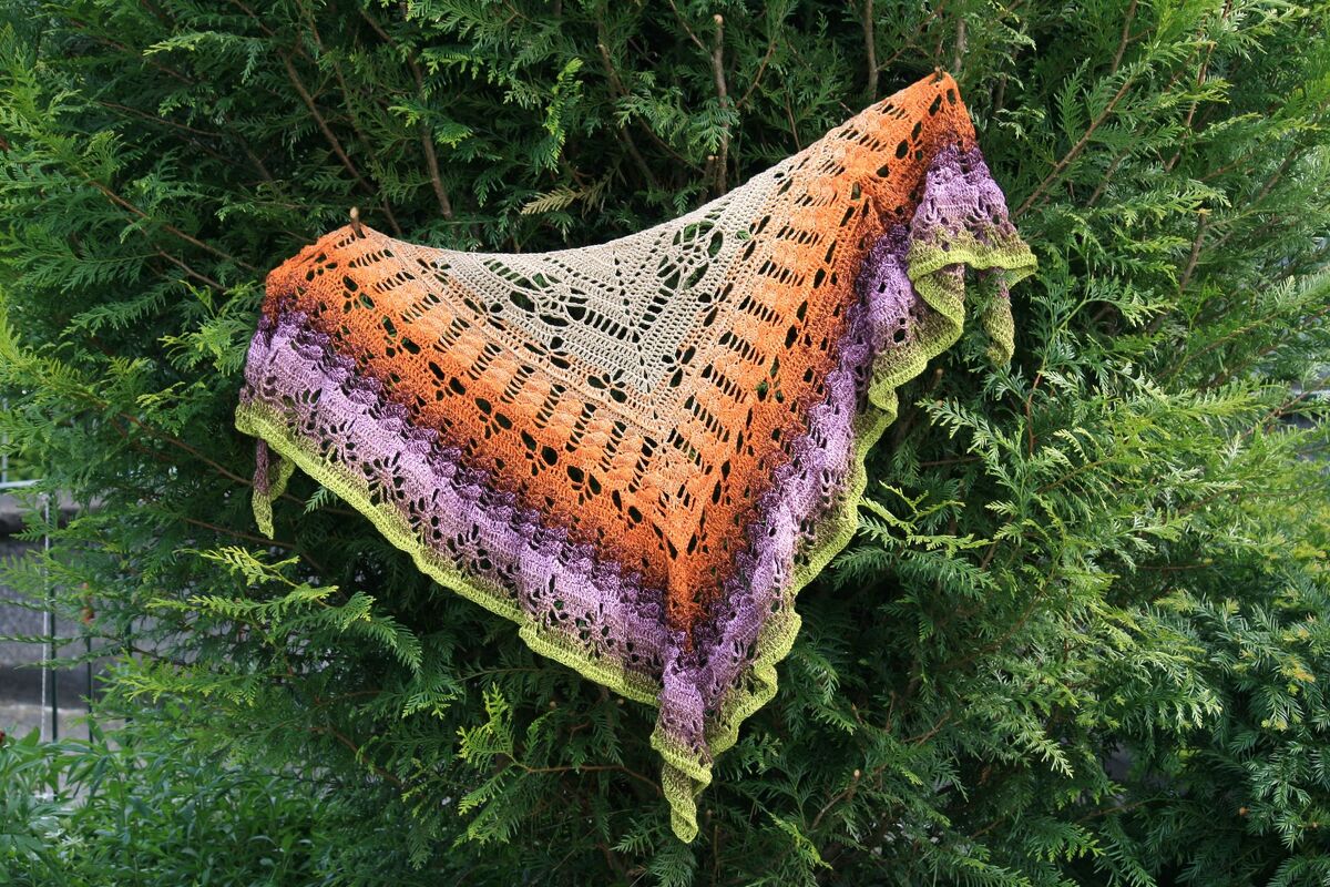 Gehäkeltes Dreieckstuch mit Farbverlauf in Beige, Orange, Lila und Grün, an einer immergrünen Hecke drapiert.