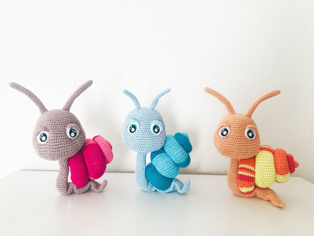 Drei gehäkelte Amigurumi‑Schnecken in Grau‑Pink, Hellblau und Orange‑Gelb vor weißem Hintergrund