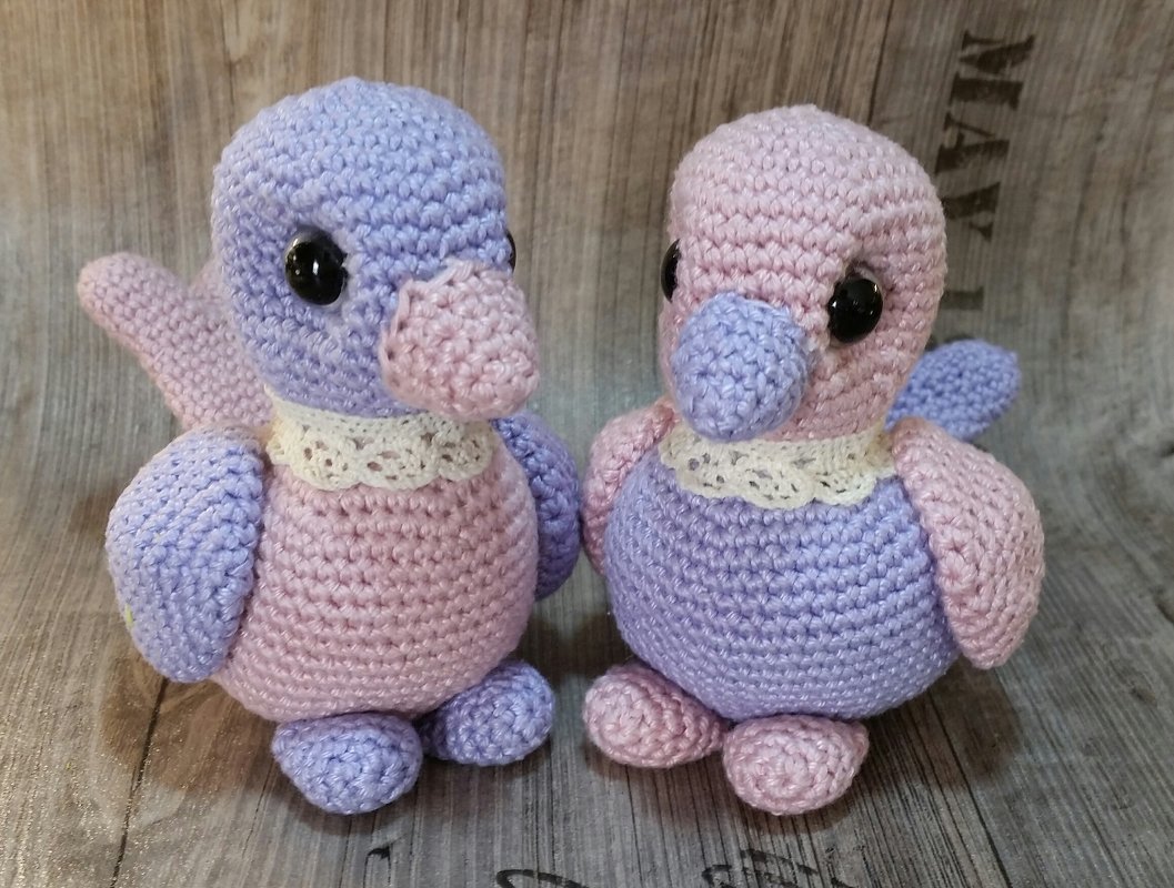 Zwei Amigurumi-Vögel aus Wolle, lila und rosa, mit weißen Kragen auf Holzoberfläche