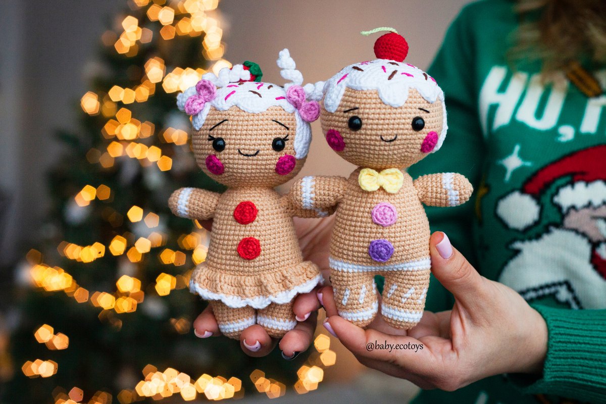 Two crocheted gingerbread amigurumi dolls with icing hair, button decorations and smiling faces held in hands with Christmas lights bokeh behind.