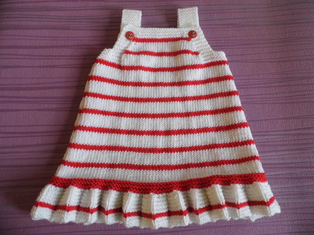 Gestricktes Baby-Trägerkleid rot-weiß gestreift mit Trägern, roten Knöpfen und Rüschensaum auf violettem Stoff.