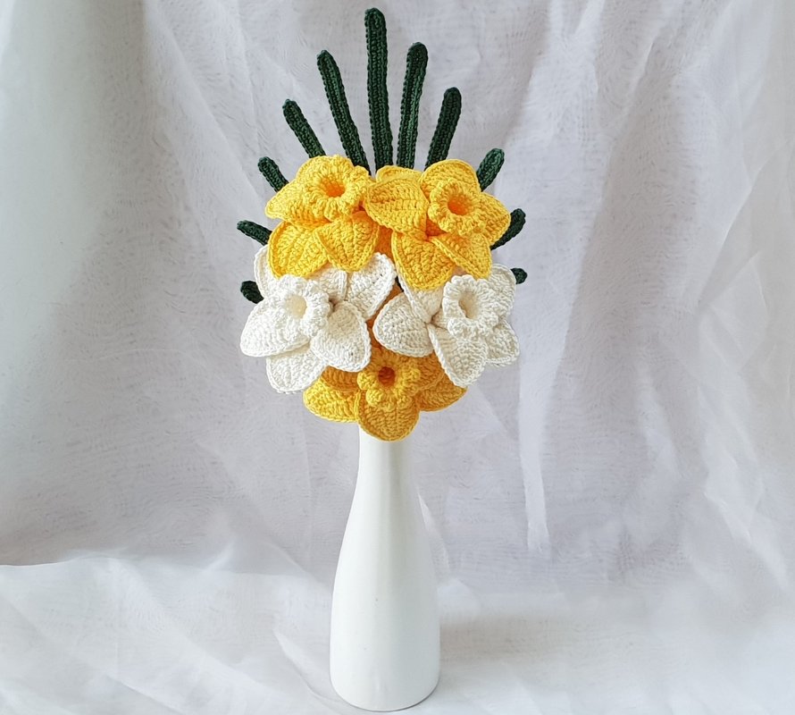 Gehäkelter Narzissenstrauß mit gelben und weißen Blüten und grünen Stielen in einer weißen Vase vor hellem Hintergrund