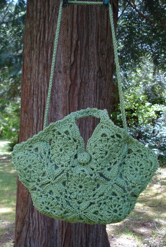 Light green crocheted bag with granny-square motifs hanging on a tree.
