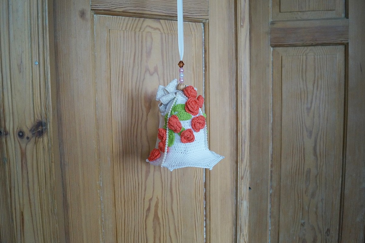 Weißes gehäkeltes Säckchen mit roten Rosen, grünen Blättern und Perlen an einem Band vor einer Holztür