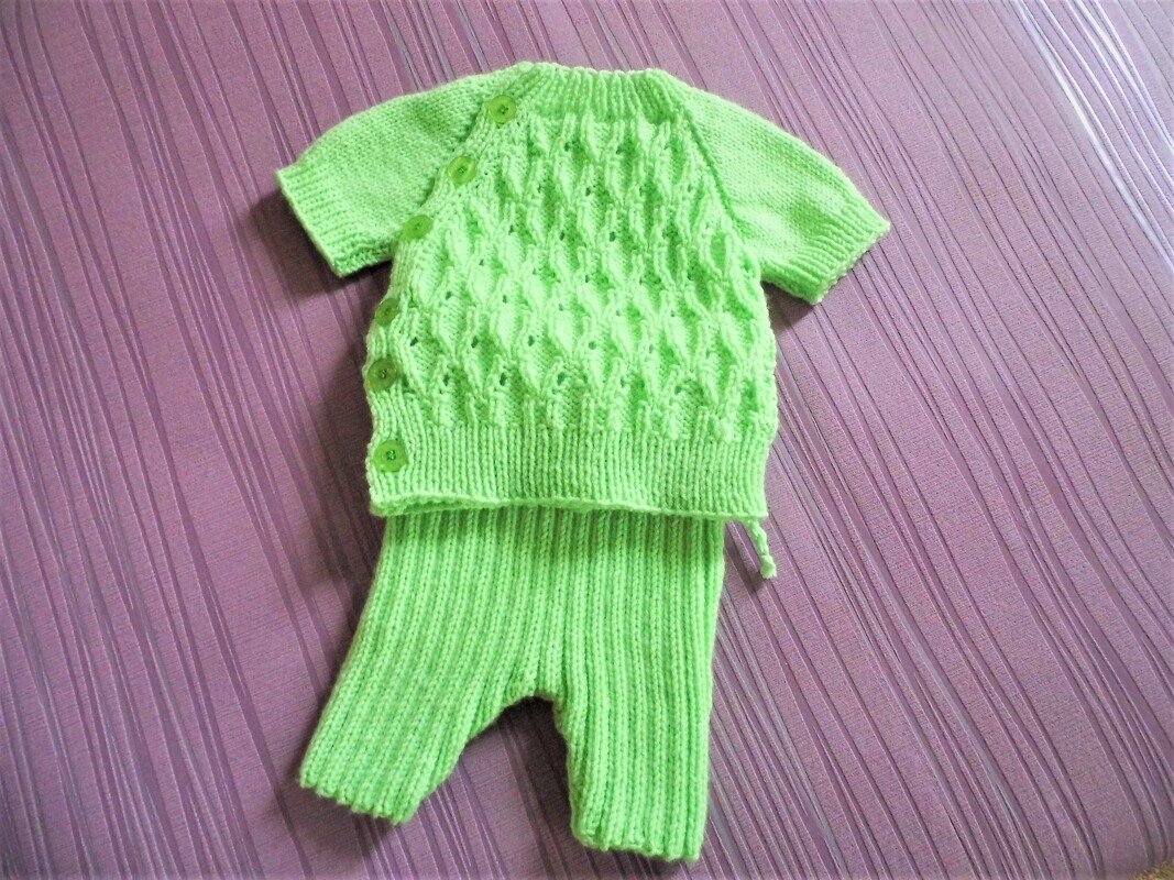 Zweiteiliges gestricktes Baby-Set in Lindgrün mit kurzärmligem Oberteil und Rippstrickhose auf violettem Untergrund.