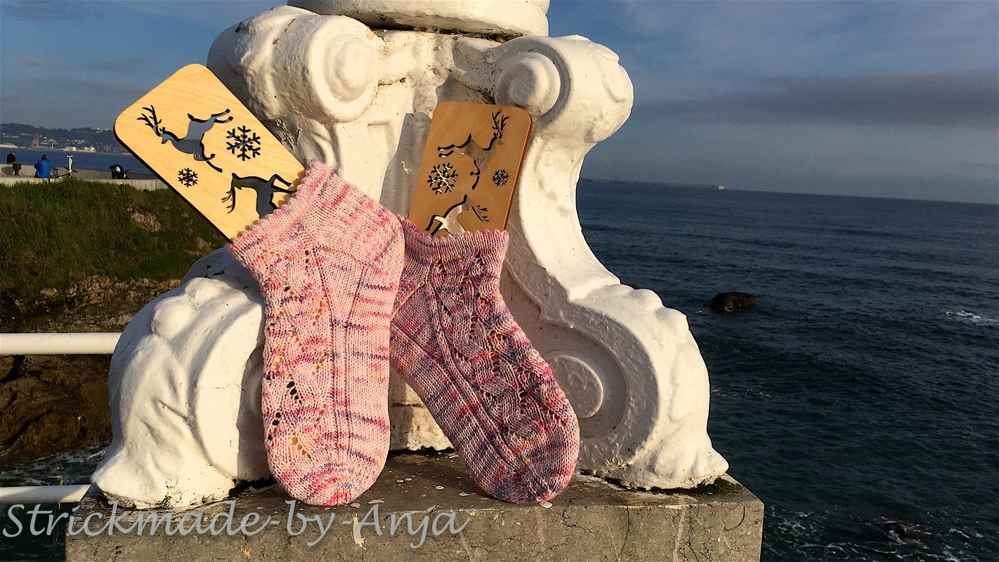 Zwei gestrickte Damensocken in Rosa- und Violetttönen mit Zopf- und Lochmuster auf weißem Steinsims vor dem Meer, mit dekorativen Holzschildern im Hintergrund
