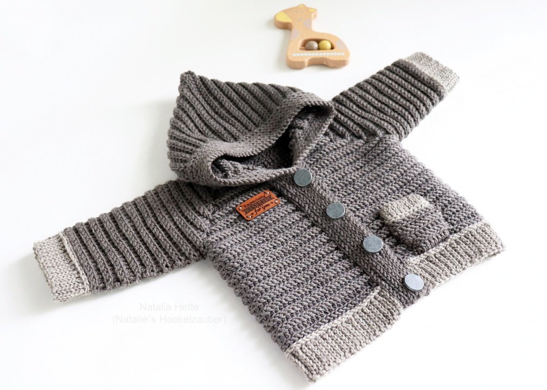 Gehäkelte graue Kinderjacke mit Kapuze, Rippenbündchen und Knopfleiste auf weißem Hintergrund, daneben ein Holzspielzeug.