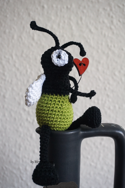 Gehäkeltes Amigurumi Glühwürmchen in Schwarz und Grün mit rotem Herz auf Tassenrand