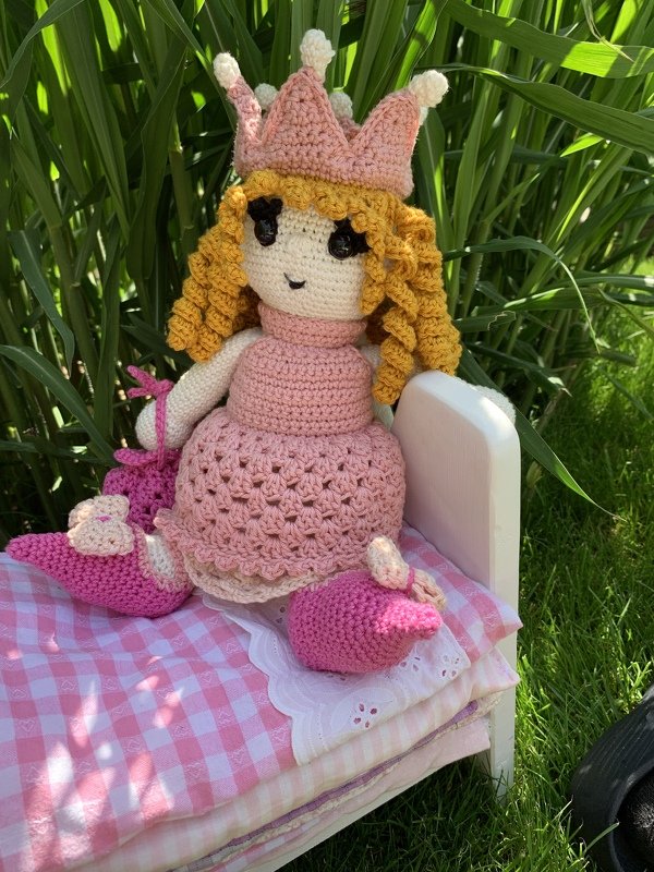 Gehäkelte Amigurumi-Puppe mit strukturiertem rosa Kleid, Krone und gehäkelten Schuhen auf einem kleinen Bett im Gras