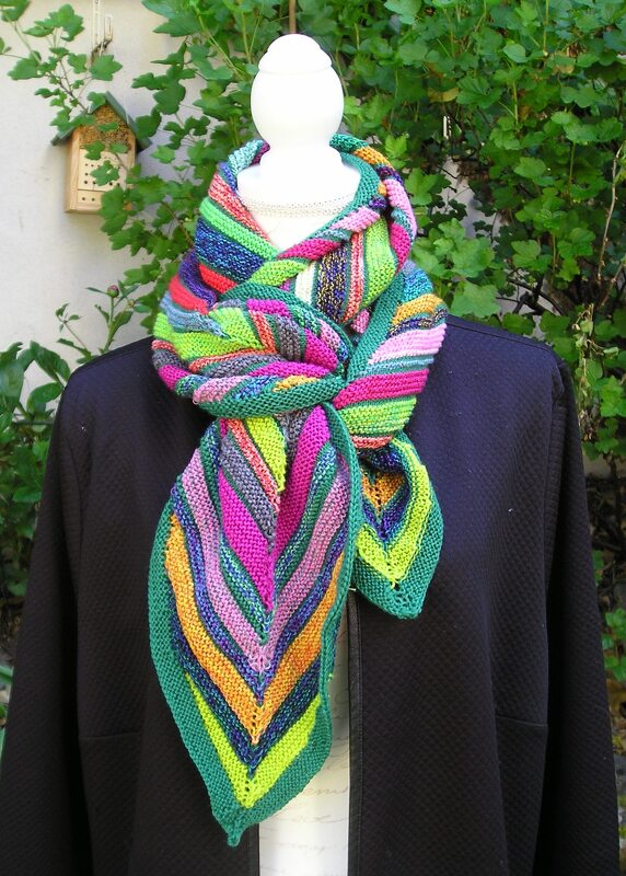 Mehrfarbiger gestrickter Schal mit Chevron-Zickzackmuster auf einer weißen Schneiderpuppe vor grünem Laub.