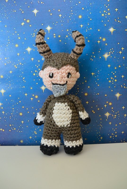 Gehäkeltes Amigurumi in braun-weißem Ziegenkostüm mit gestreiften Hörnern und grauem Bart vor blauem Sternenhintergrund