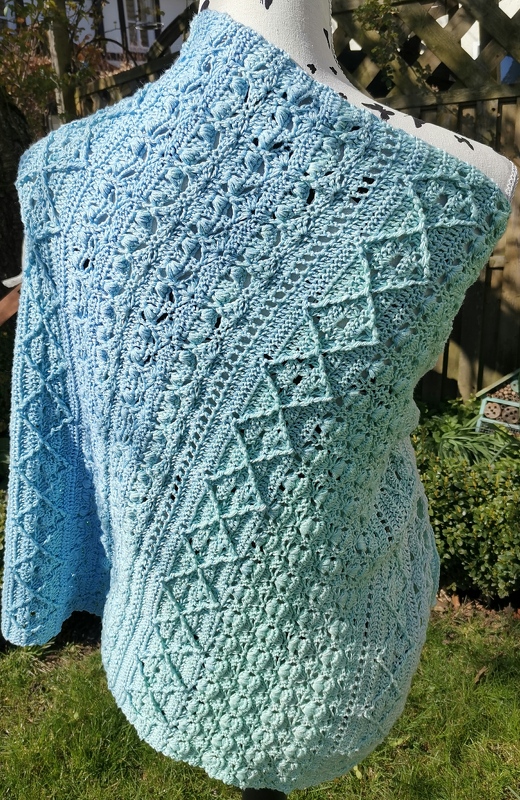 Gehäkeltes Schultertuch in Hellblau bis Mintgrün mit Noppen- und Rautenmuster, auf einer Schneiderpuppe drapiert.