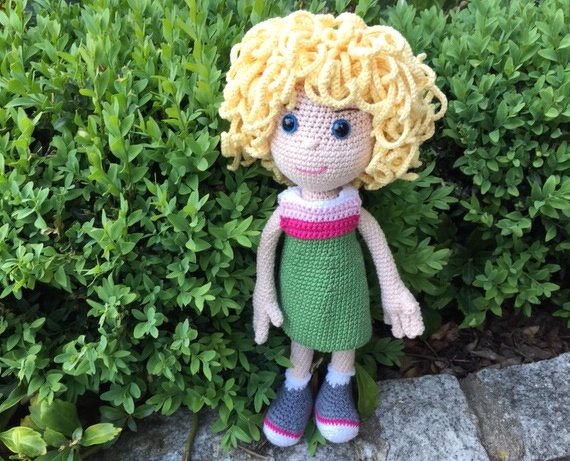 Gehäkelte Amigurumi‑Puppe mit lockigen blonden Haaren, grünem Kleid und grauen Schuhen vor Buschwerk.