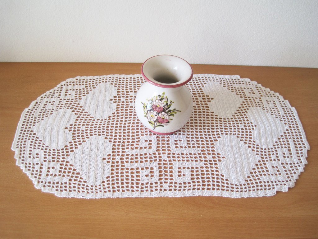 Weißes, ovales Häkeldeckchen mit herzförmigen Mustern auf einem Holztisch, darauf eine kleine Blumenvase